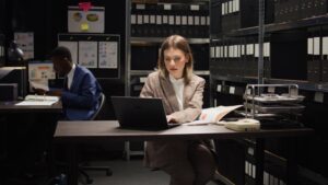 In office, detective inspector and male police officer conduct an investigation, expertly analyzing files and evidence. Portrait of caucasian law officer examining information on paper with laptop.